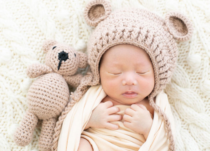 ニューボーン：クマ耳の付いたニット帽をかぶりクマのぬいぐるみと並んで眠る赤ちゃん
