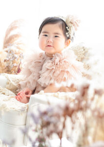 ベビー・バースデー：ドライフラワーに囲まれ微笑むくすみピンクのチュールフリルドレスを着た女の子