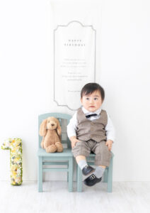 ベビー・バースデー：犬のぬいぐるみと並んで蝶ネクタイとベストを来て水色の椅子に腰かけた男の子