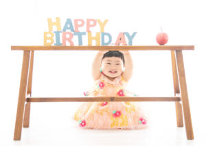 ベビー・バースデー：お花がついたピンクとオレンジのドレスを着て笑顔でこちらを見つめる女の子