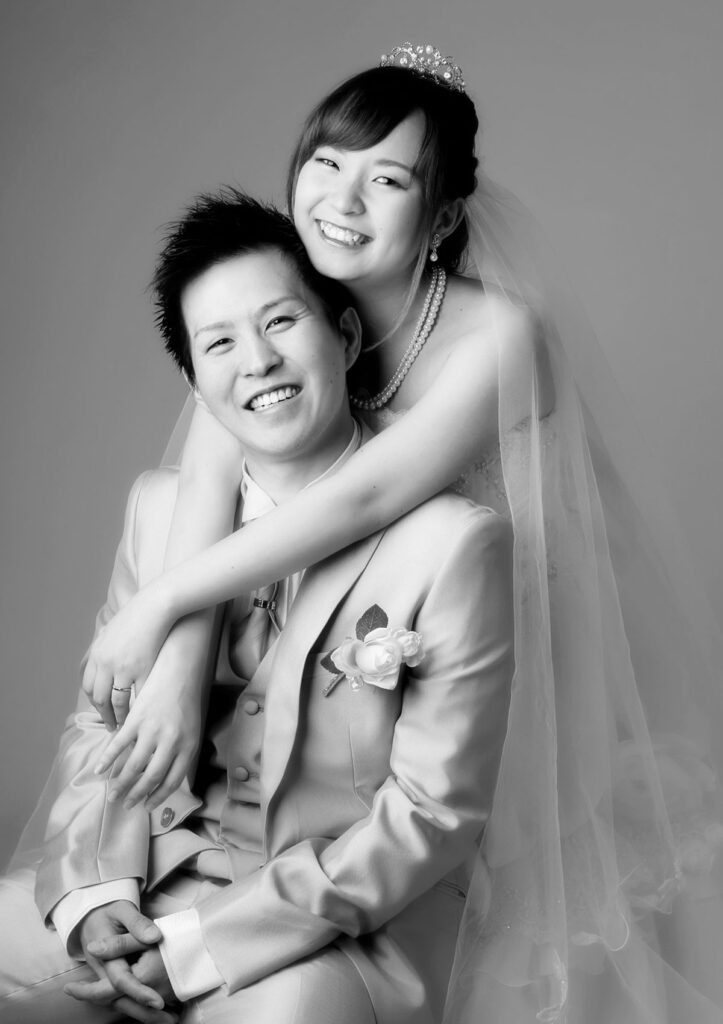 ウェディング：タキシードを着た新郎をウェディングドレスを着た新婦がバックハグをして仲良く笑う様子