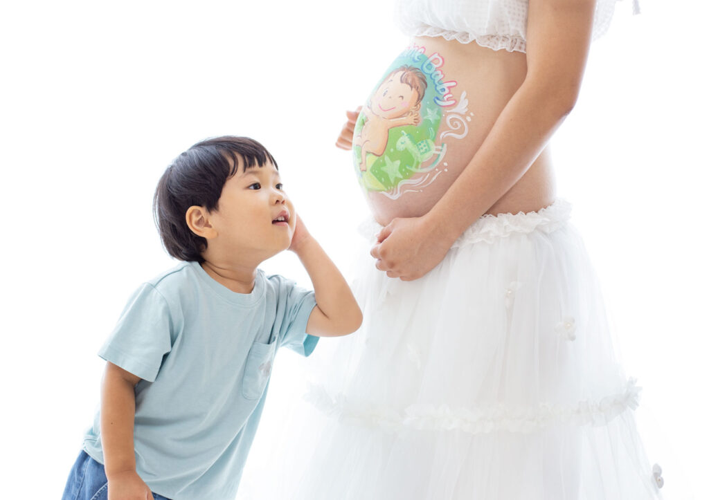 マタニティ：白いマタニティドレスでお腹にベリーペイントを施したママのお腹に、顔を近づけて耳を澄ませる男の子のツーショット