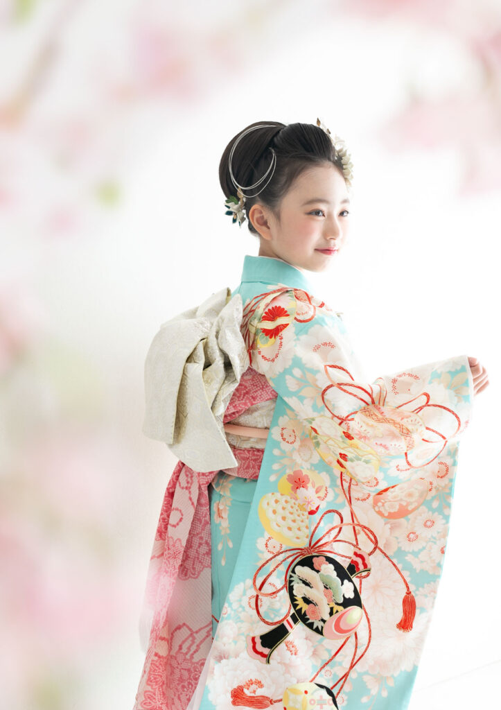U2025-7model_753 七五三:水色で古典柄の優美な着物を身にまとい、桜の花越しに振り返る7歳の女の子