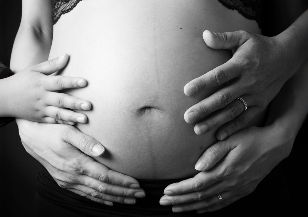 マタニティ：大きなおなかにパパ、ママ、子どもの三人の手を重ねたお腹アップのモノクロフォト