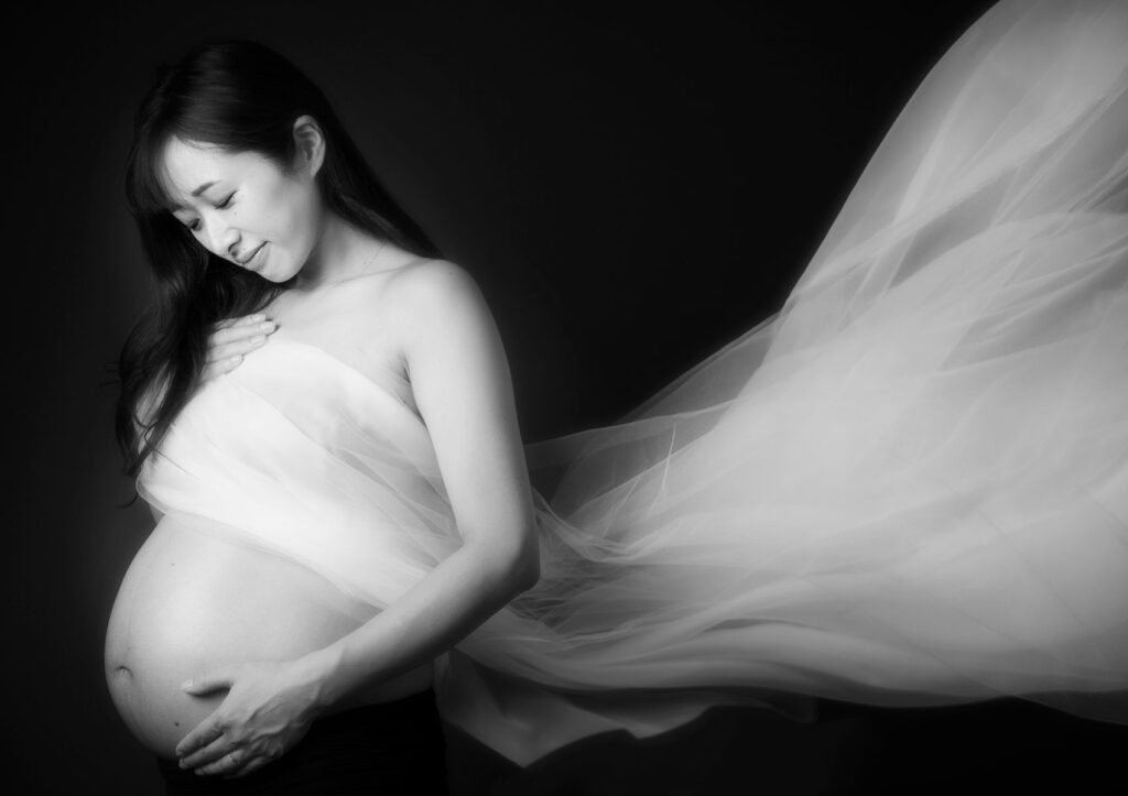 マタニティ：大きなお腹を愛おしそうに見つめるママ、胸元から背中にかけてドレープが優雅にたなびく神秘的なモノクロフォト