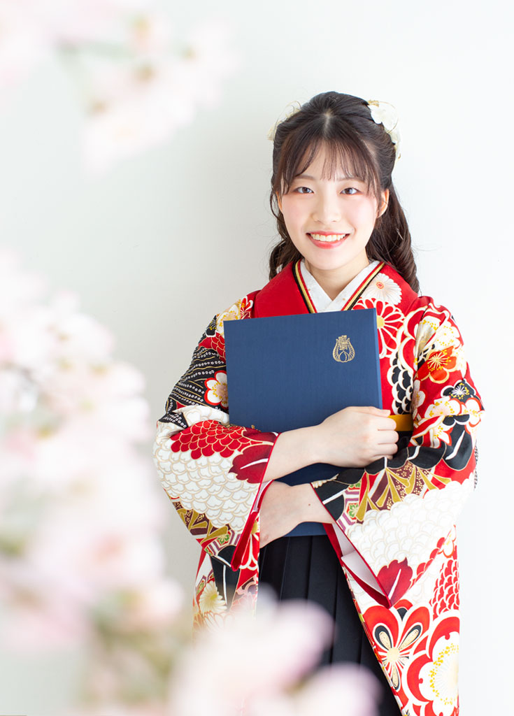 卒業袴：手前に映る桜とともに、両手で卒業証書を大事に抱えた女性