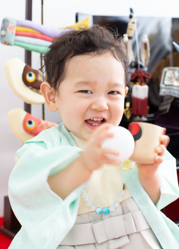 お節句：着物を着て、鯉のぼりや兜人形を背におもちゃで遊ぶ男の子