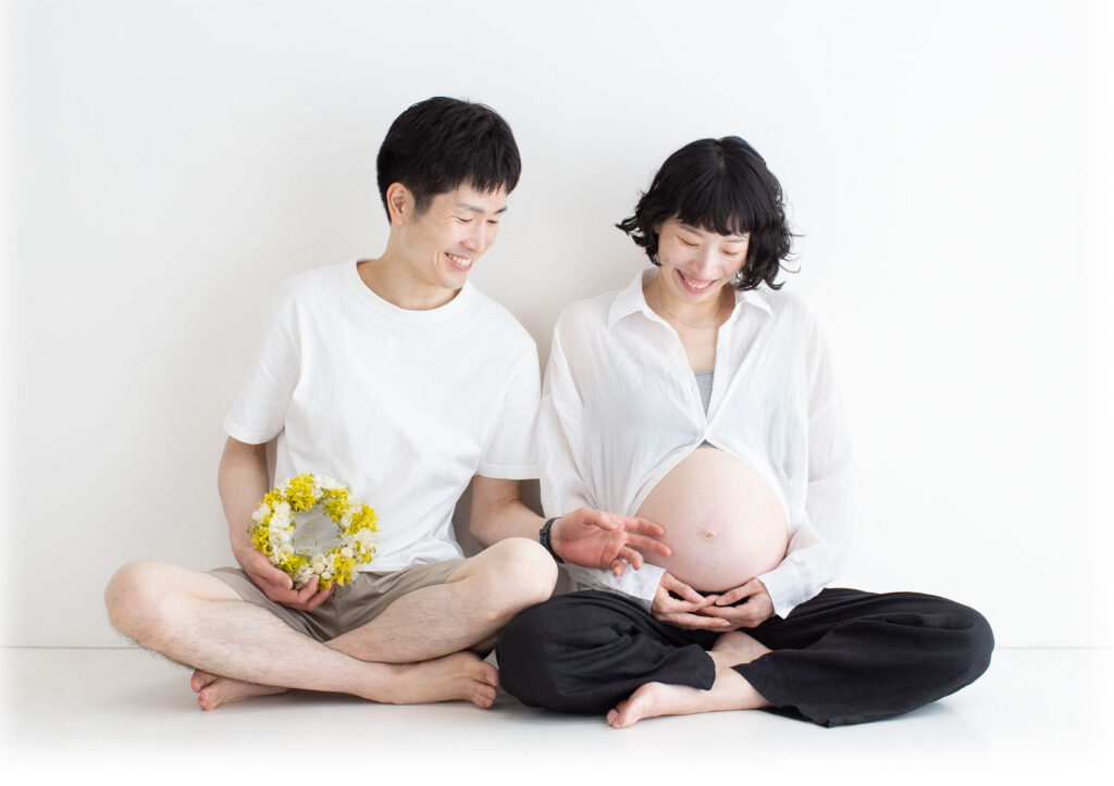 マタニティ：ナチュラルなシャツ姿でお腹を見つめながら微笑んでいる、胡坐をかいたパパとママ