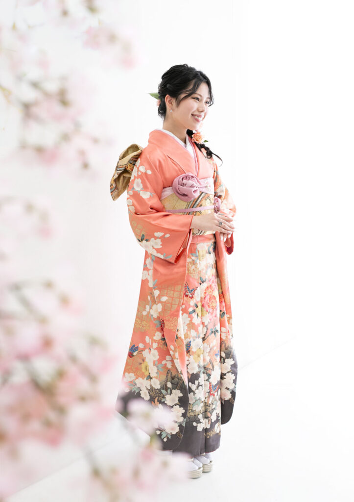 成人式：コーラルカラーの振袖を着て、軽やかに微笑む女性と桜の花