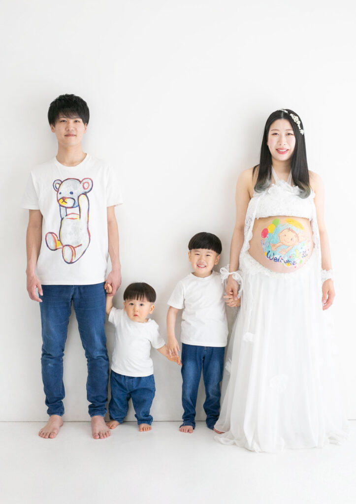 マタニティ：白いマタニティドレスをまといお腹にベリーペイントを施したママと、白シャツ・デニムのリンクコーデをした男の子2人とパパが横並びに手をつないだ家族写真