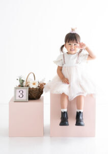 ベビー・バースデー：ピンクのボックスに腰かけて目元でピースをする女の子