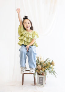 ベビー・バースデー：椅子に座って右手を大きく上げる笑顔の女の子