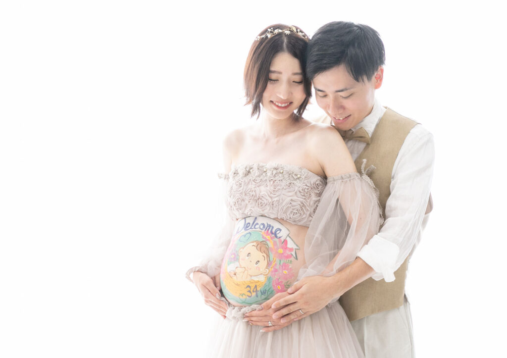 マタニティ：グレージュのチュールドレスを着てお腹にベリーペイントを施したママに、バックハグで寄り添うパパのツーショット