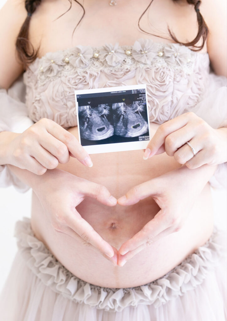 マタニティ：グレージュのチュールドレスを着てエコー写真を手に持つママと、ママのおへその前でハートマークを手でつくるパパとの、お腹のアップの写真