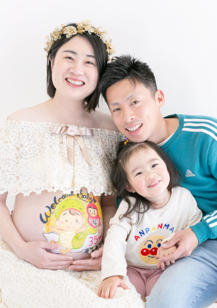 マタニティ：ナチュラルなレースのドレスを着てベリーペイントをお腹にあしらったママに寄り添うパパと、お姉ちゃんの家族写真