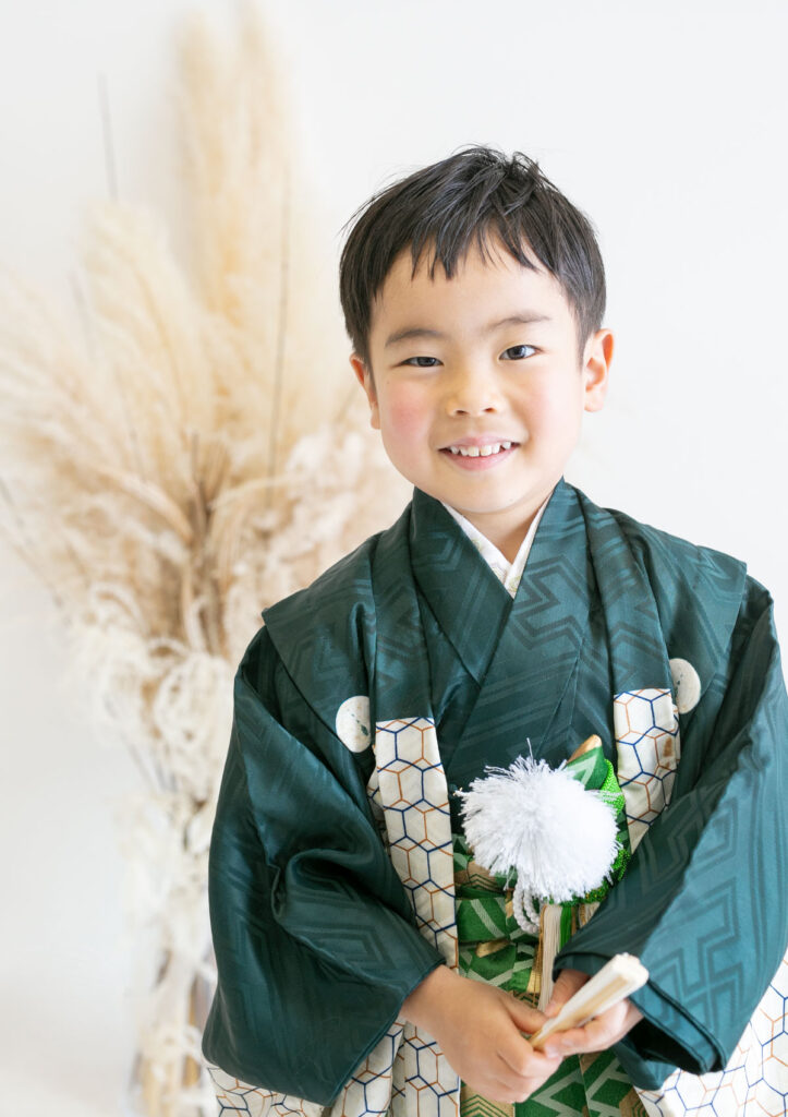 U250127_753 七五三:深い緑の羽織袴に扇子を持ち、やさしく微笑む5歳の男の子