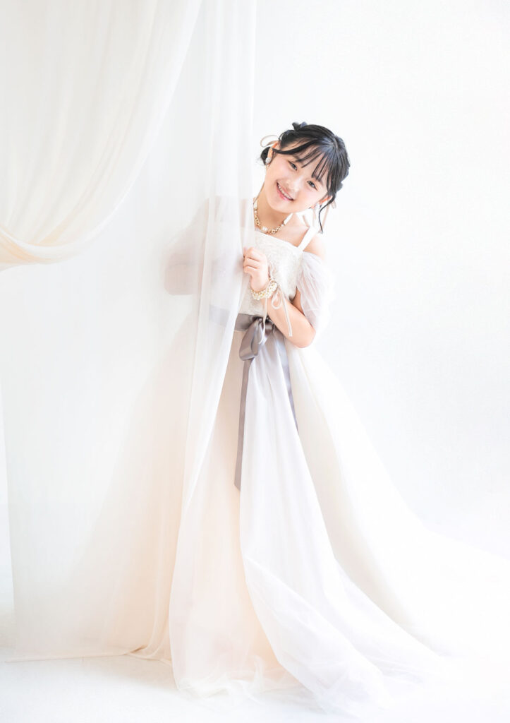 ハーフ成人：少し透けているドレープから顔をのぞかせるホワイトのドレスを着た10才の女の子