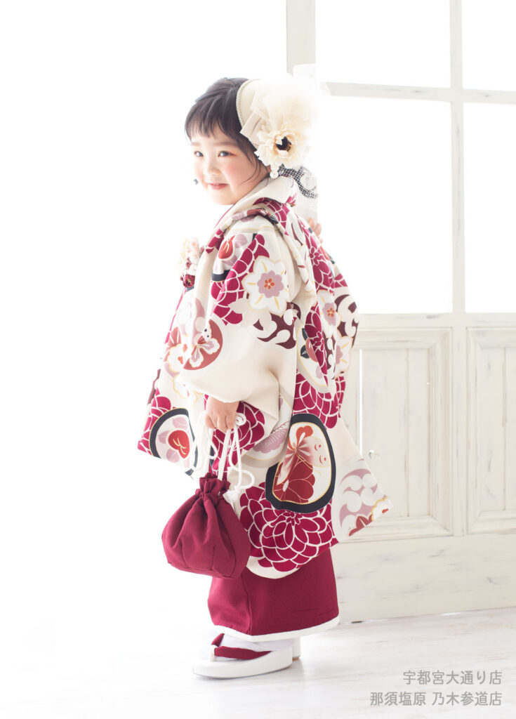 七五三３歳着物：エンジ色に大きく菊の花が描かれた、大きな髪飾りを着けた３才の女の子。