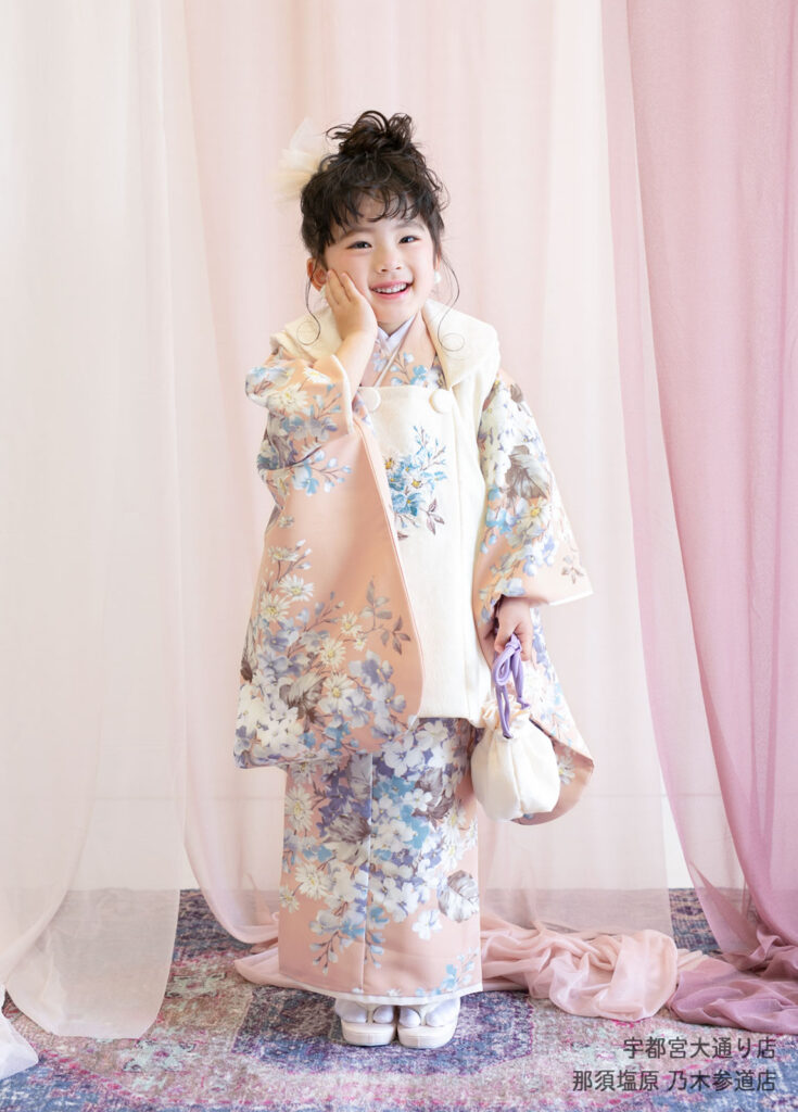 七五三３歳着物：くすみピンクの花柄の着物を着て頬に手を当てる女の子。
