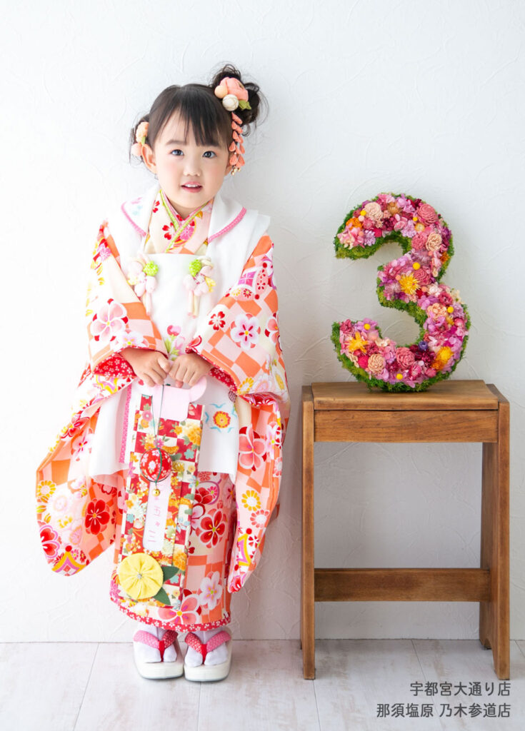 七五三３歳着物：オレンジの華やかな柄の着物を着た千歳あめを持った３才の女の子。