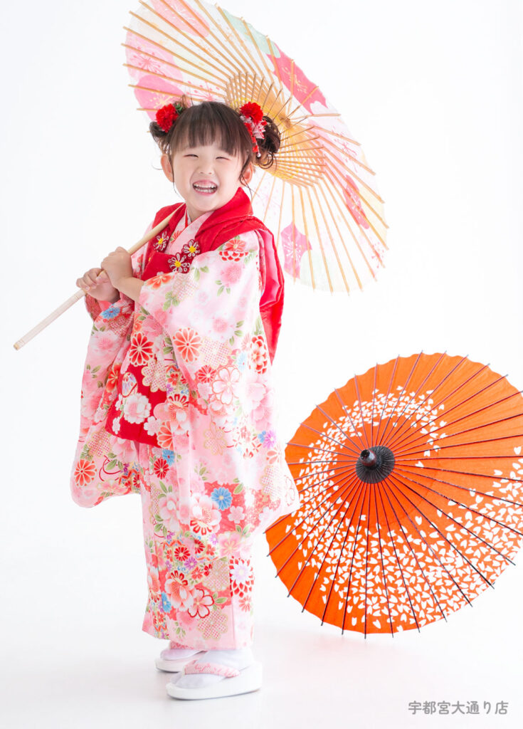 七五三３歳着物：ピンクの花柄の着物を着て和傘をもって笑う３才の女の子。