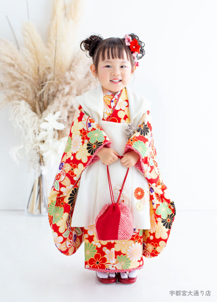 七五三３歳着物：赤い和柄の着物に白い被布を合わせたツインアップスタイルの３才の女の子。