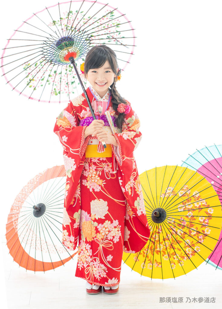 七五三７歳着物：真っ赤な着物に和傘を持ち、花の髪飾りを着けた７才の女の子。