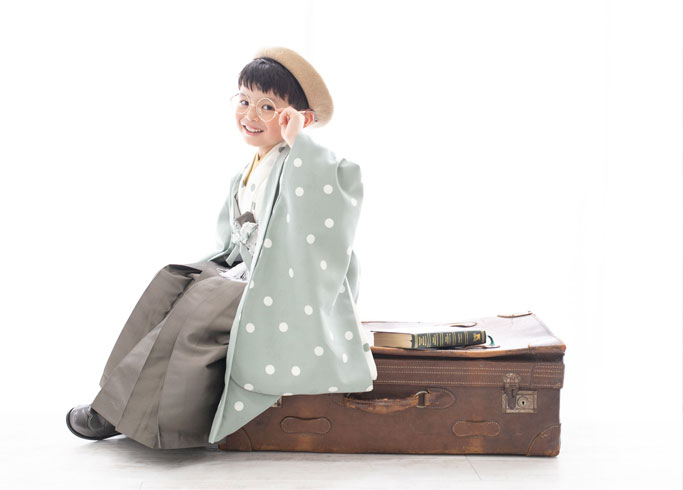 七五三：くすみグリーンの水玉柄の羽織袴にベレー帽、眼鏡を身に着け、大きなトランクケースに腰かけた5歳の男の子