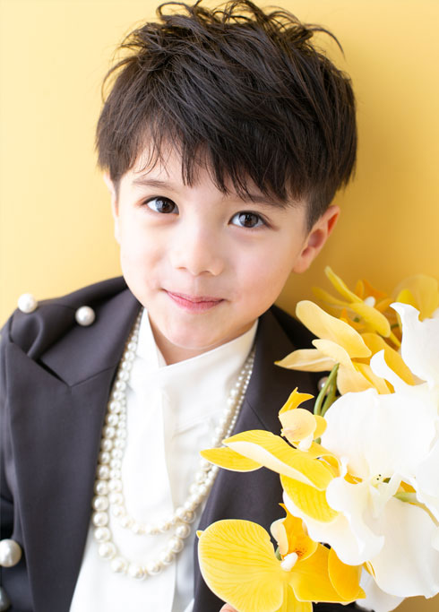 七五三：パールがあしらわれたジャケットスーツとパールのネックレスを身に着け、胡蝶蘭を片手に微笑む5歳の男の子
