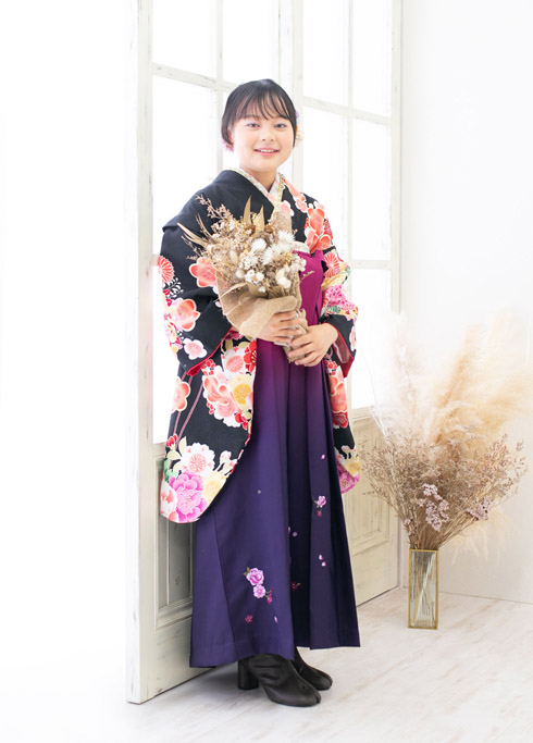 ハーフ成人：黒い古典柄の二尺袖に紫・ピンクのグラデーションの袴を着て花束を持つ、10歳の女の子