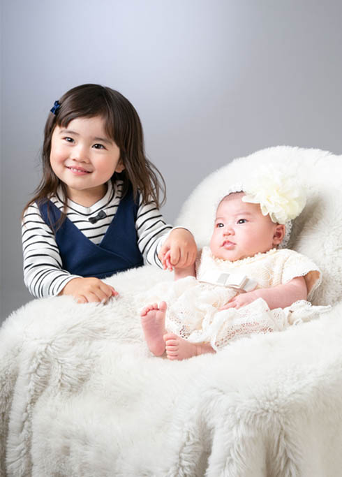 お宮参り：お姉ちゃんと手をつなぐ、レースのベビードレスを着た女の子の赤ちゃん
