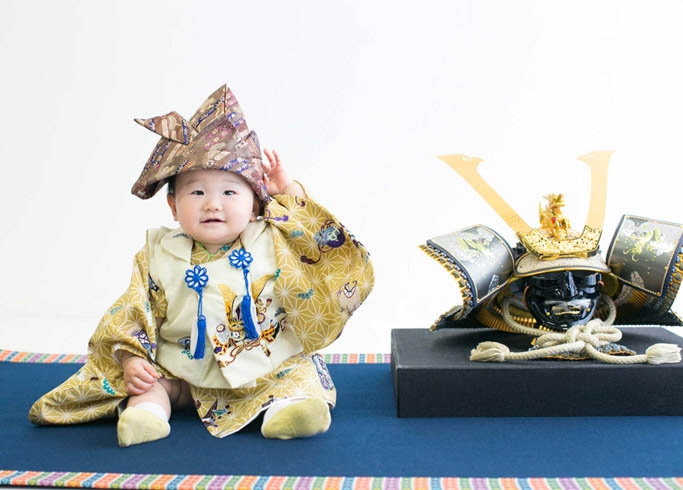 端午の節句：兜飾りと並んで兜の帽子をかぶる男の子の赤ちゃん。