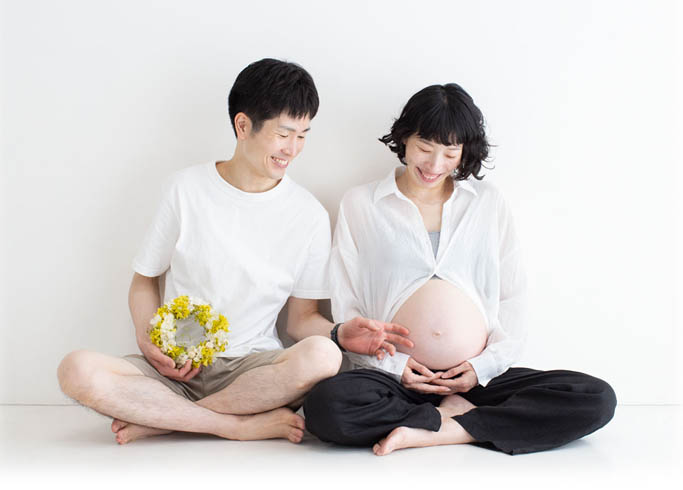 マタニティ：胡坐で大きなお腹を服から覗かせ、やさしく手を伸ばすパパとママとのツーショット