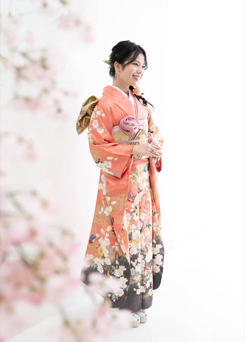 成人：珊瑚のようなコーラルピンクの振袖を着て桜の木越しにたたずむ、20歳の女性