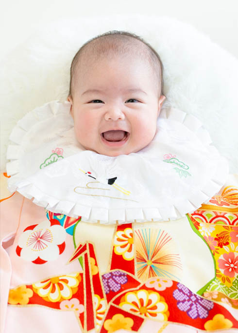 お宮参り：ピンク色で古典柄の産着を着て、口を開けにっこり笑う女の子の赤ちゃん