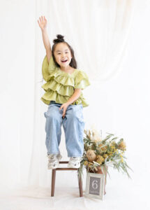 ベビー・バースデー：ライトグリーンのフリルトップスを着て元気に手を挙げて返事をする6歳の女の子。