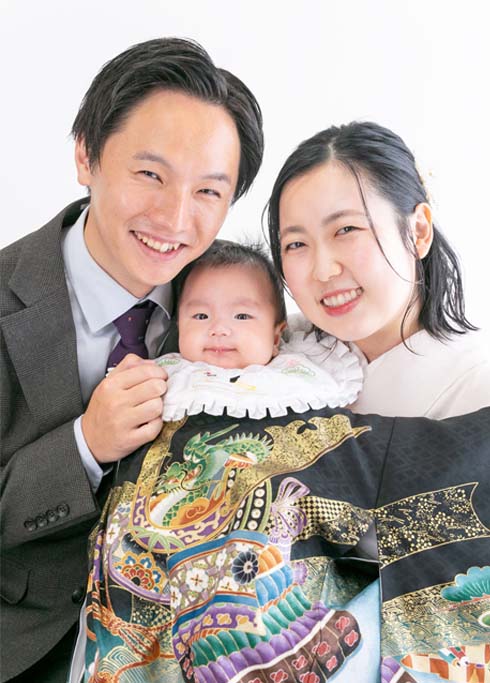 お宮参り：黒い古典柄の産着を着て、スーツを着たパパと着物を着たママにハグされる男の子の赤ちゃん