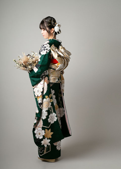 成人：深い緑色で古典的な柄の振袖を着て花束を持つ、二十歳の女性