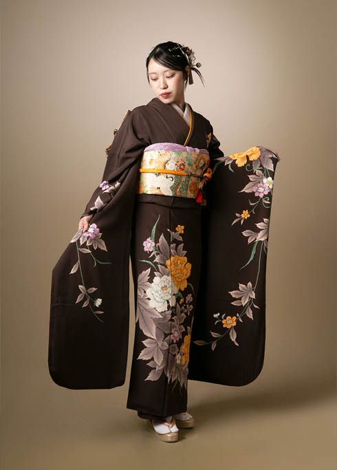 成人：深い茶色にシンプルかつ大胆な花柄の振袖を着て、袖を広げて立つ20歳の女性