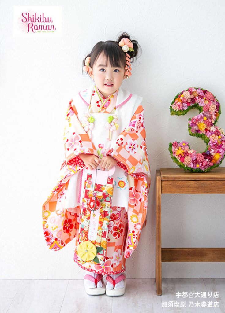 七五三３歳：オレンジ色の市松模様を基調として花柄をあしらった着物に白の被布を着た3才の女の子。