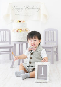 ベビー・バースデー：オシャレなスーツを着てケーキの前に座る笑顔の1才の男の子。